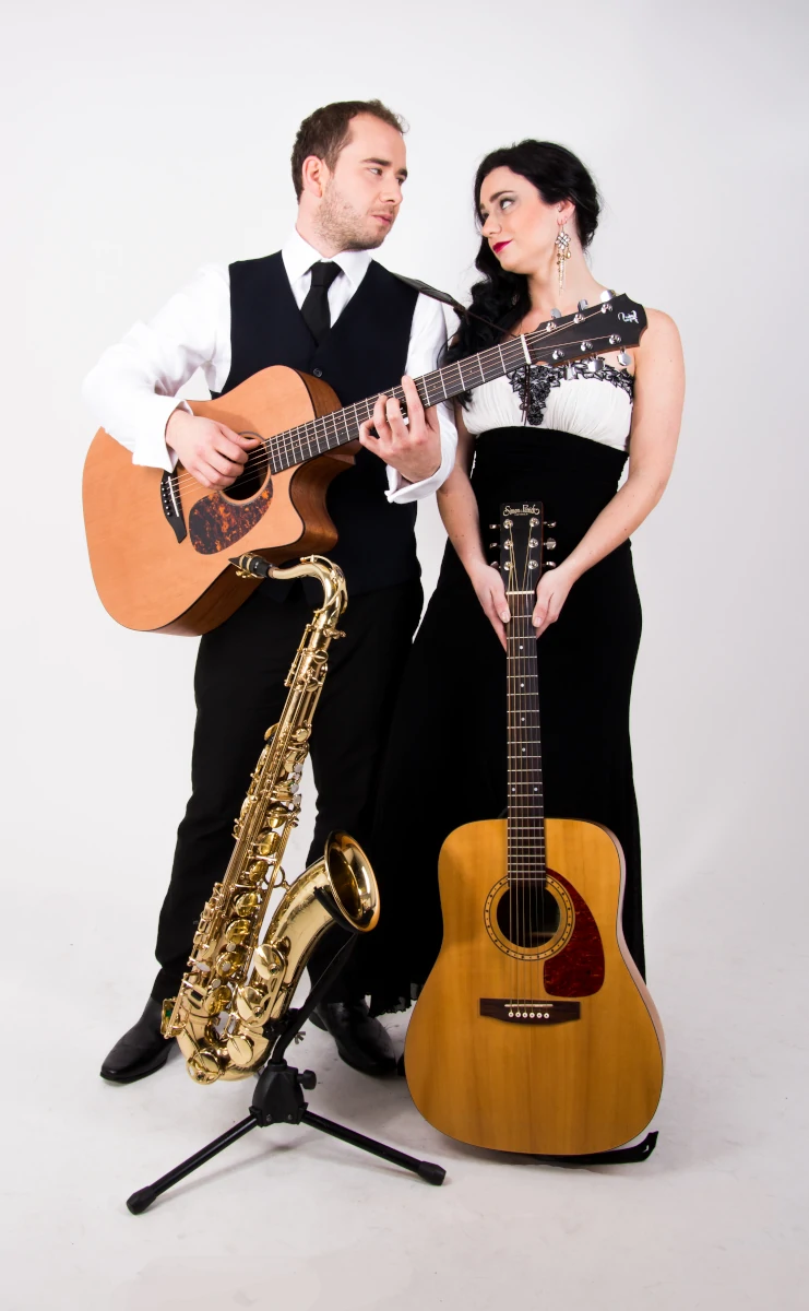 A photo of Double Malt Duo in smart attire and looking at each other, with 2 guitars and a saxophone.
